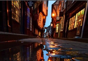 A narrow street with buildings on either side, reflecting lights on the wet ground.