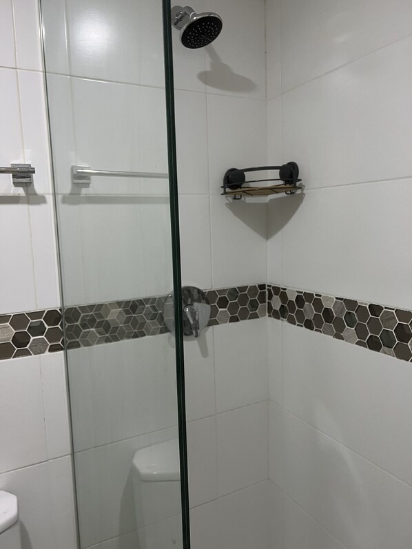 A glass-enclosed shower with a handheld showerhead, a towel rack, and a shelf with toiletries. - Bayfront Apartment & villa (Portmore)