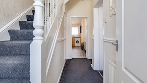 Un escalier avec des balustrades blanches menant à un palier, un couloir avec une porte sur la droite, et, au fond, on aperçoit une partie de la cuisine.