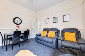 A modern living room with a dining area, featuring a sofa set, a dining table with chairs, and a wall clock.