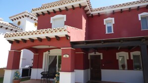 Ein zweistöckiges Gebäude mit roter Fassade, weißem Zierdekor und einem Ziegeldach. Eine Schilder mit der Nummer 56, eine Tür und ein kleiner Patio mit Tisch und Stühlen sind zu sehen.
