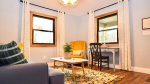 A living room with a sofa, a small table with a plant, a chair, and a desk with a chair. There are two windows with curtains, a ceiling light, and a wooden floor. - Heartwood♥ Cabin loft w/private HotTub, Hiking, Fishing, & Ziplines on 66 acres! (Floyd)