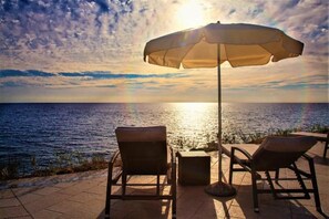 Un patio frente al mar con dos sillones de descanso y una mesa bajo un gran paraguas, con vista al océano.