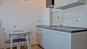 Una pequeña kitchenette con cubierta blanca, un lavamanos y una estufa. Hay una mesa de comedor con dos sillas y un reloj en la pared.
