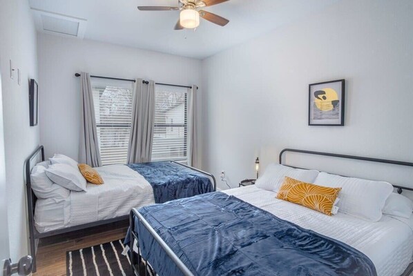 A bedroom with two beds, a ceiling fan, a framed picture on the wall, and a window with blinds. - Cutting-Edge Elegant 3BR 2.5BA Home Near Downtown! (San Antonio)