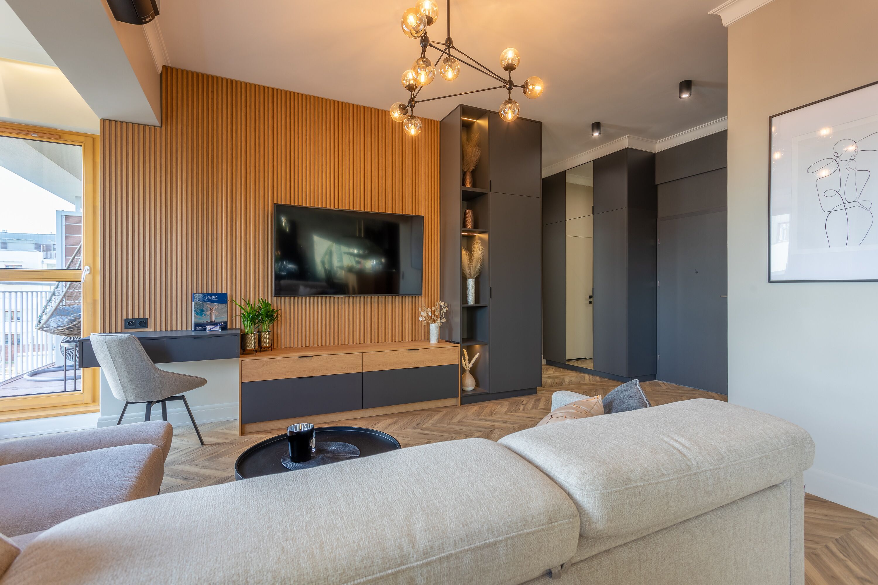 Ett modernt vardagsrum med en soffa, ett soffbord och en tv som är monterad på en väggpanel av trä. Det finns också en stol och ett litet bord med en växt.