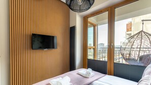 Une chambre d’hôtel moderne dotée d’un grand lit, d’un téléviseur à écran plat fixé sur un mur en panneaux de bois, et d’un balcon avec un fauteuil suspendu.