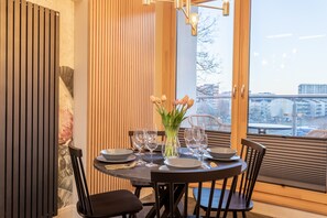 Un coin repas avec une table ronde dressée pour quatre personnes, un vase rempli de fleurs et une vue sur la ville grâce à de grandes fenêtres.