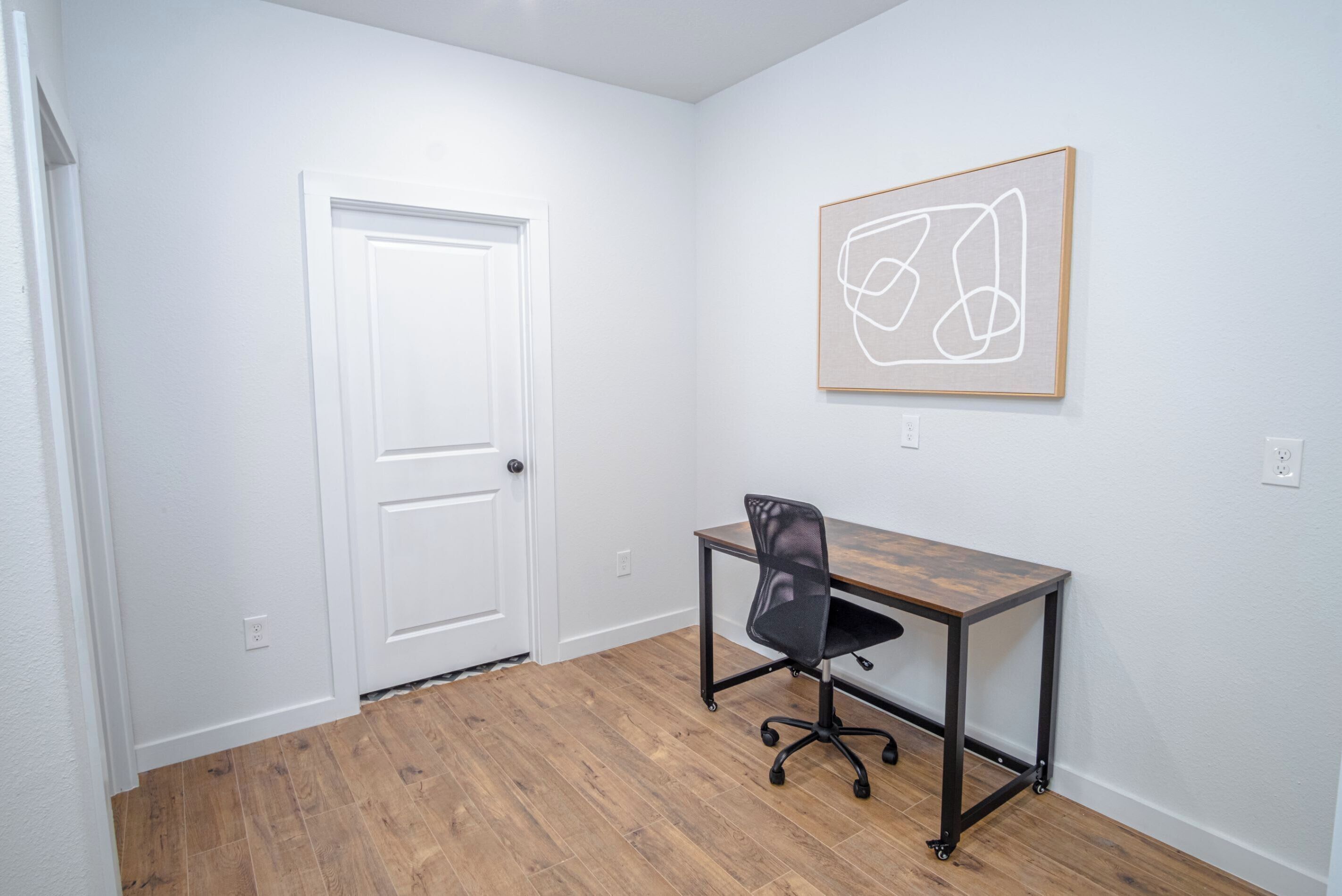 A room with a wooden floor, a white door, a black office chair, and a framed abstract artwork on the wall.