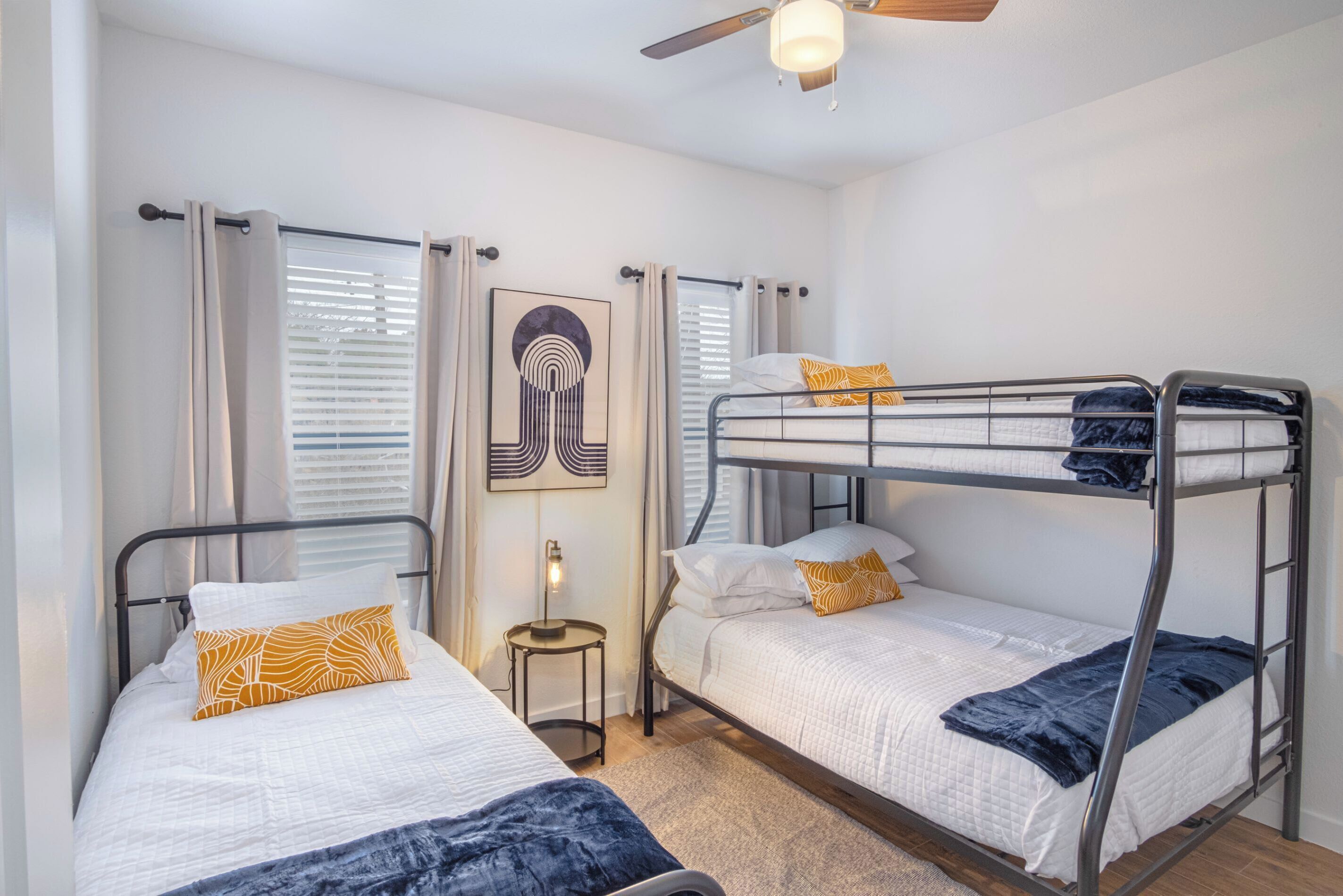 A room with two bunk beds, a ceiling fan, a small table with a lamp, and a framed artwork on the wall.