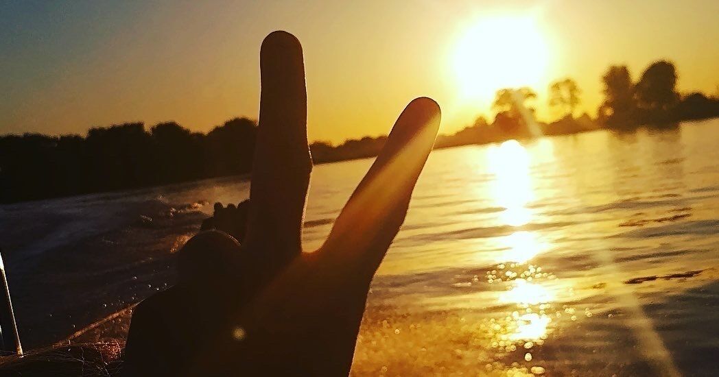Une main faisant le signe de la paix devant un coucher de soleil sur l’eau.