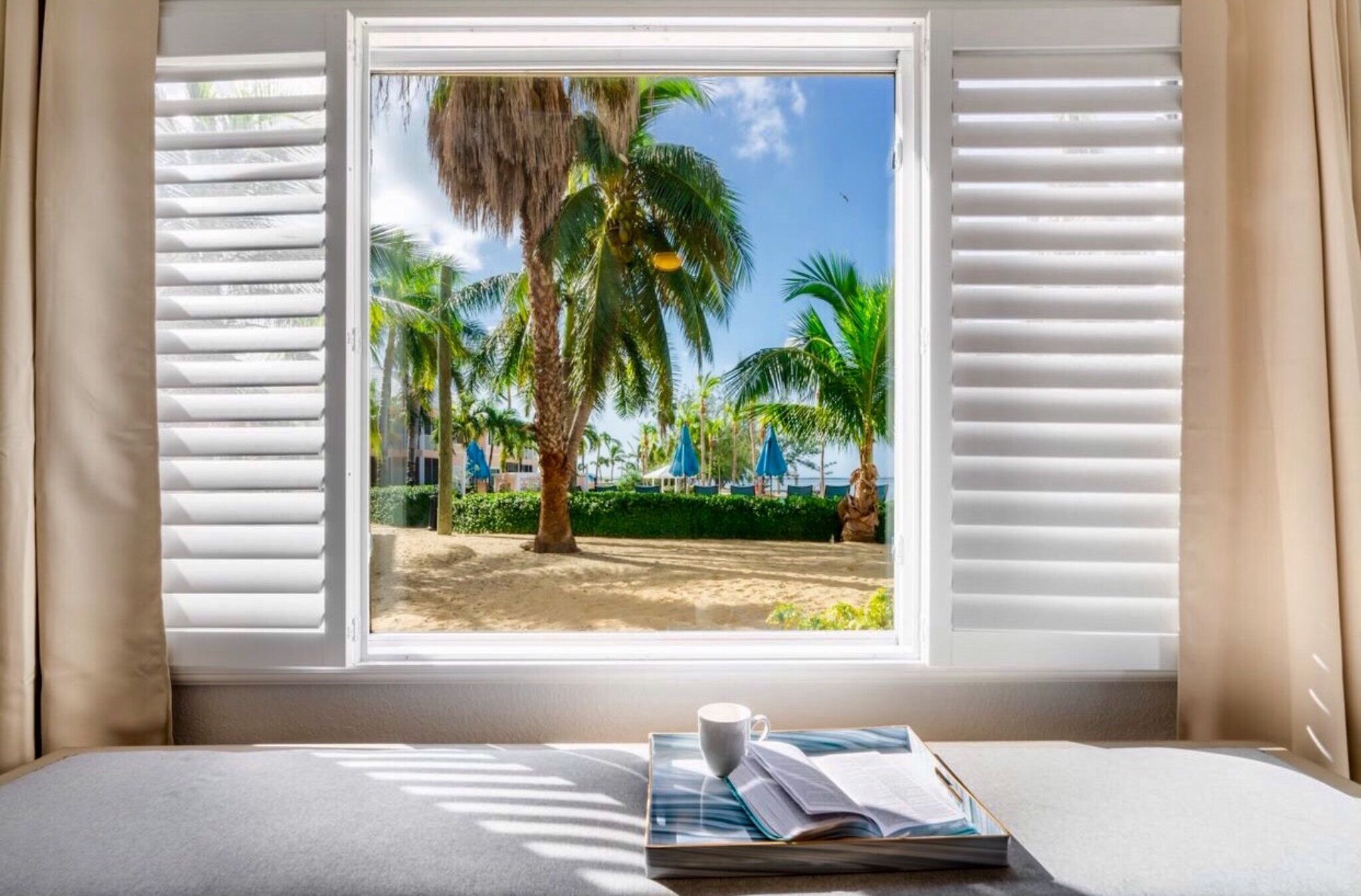 A room with a window overlooking a beach, a bed, and a table with a book and a cup.