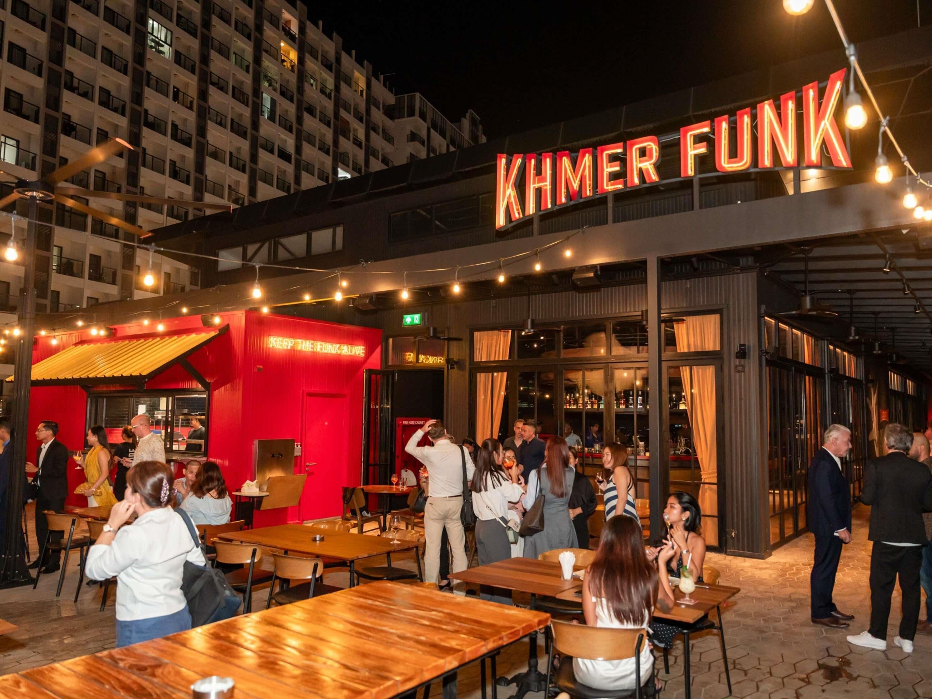 An outdoor seating area with tables and chairs, a neon sign reading 'KHMER FUNK', and a building with multiple floors in the background.