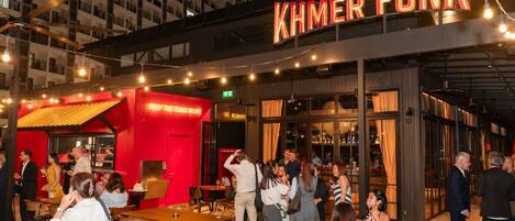 An outdoor seating area with tables and chairs, a neon sign reading 'KHMER FUNK', and a building with multiple floors in the background.