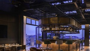 A modern bar area with a city view, bar stools, and tables with glasses. - TRIBE Phnom Penh Post Office Square (Phnom Penh)