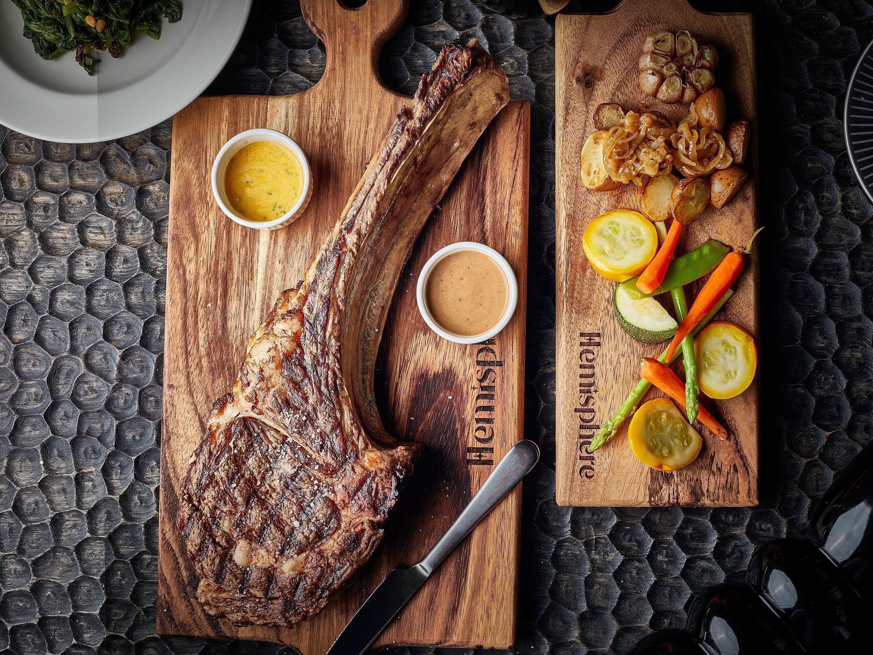 Un steak servi sur une planche en bois, accompagné de deux petits ramequins de sauce et d’un accompagnement de légumes rôtis.
