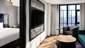 A modern hotel room with a bed, a TV mounted on a wall, a round coffee table, a sofa, and a view of the city through large windows.