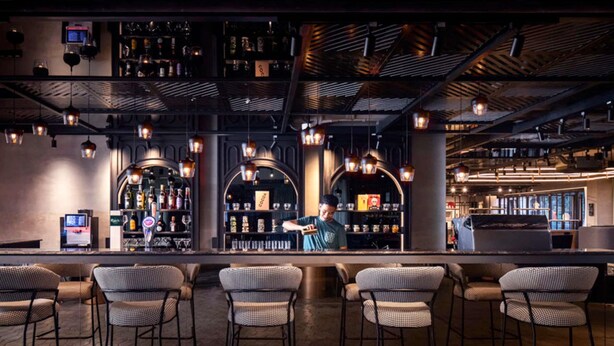 A bar area with a bartender, wicker chairs, and shelves stocked with bottles and glasses. - TRIBE Phnom Penh Post Office Square (Phnom Penh)