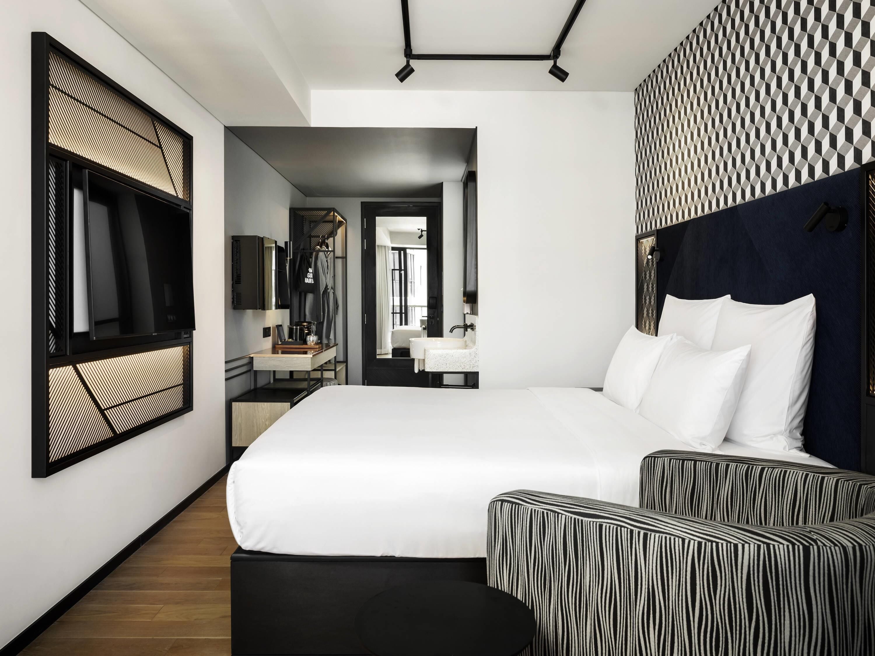 A modern hotel room with a large bed, a TV, a desk with a mirror, and a bathroom visible through an open door.