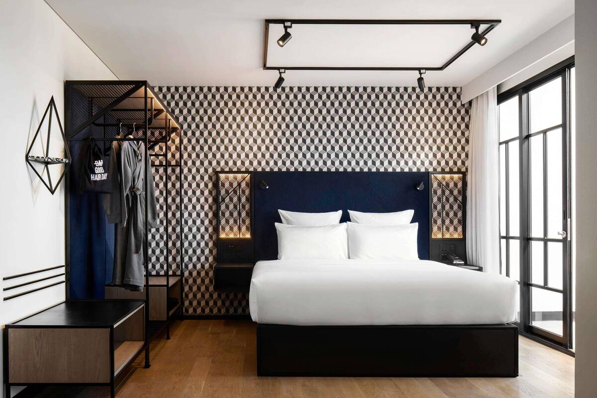 Una habitación de hotel moderna con un papel tapiz de diseño geométrico, una cama grande con ropa blanca, una mesita de noche, un escritorio y un armario.