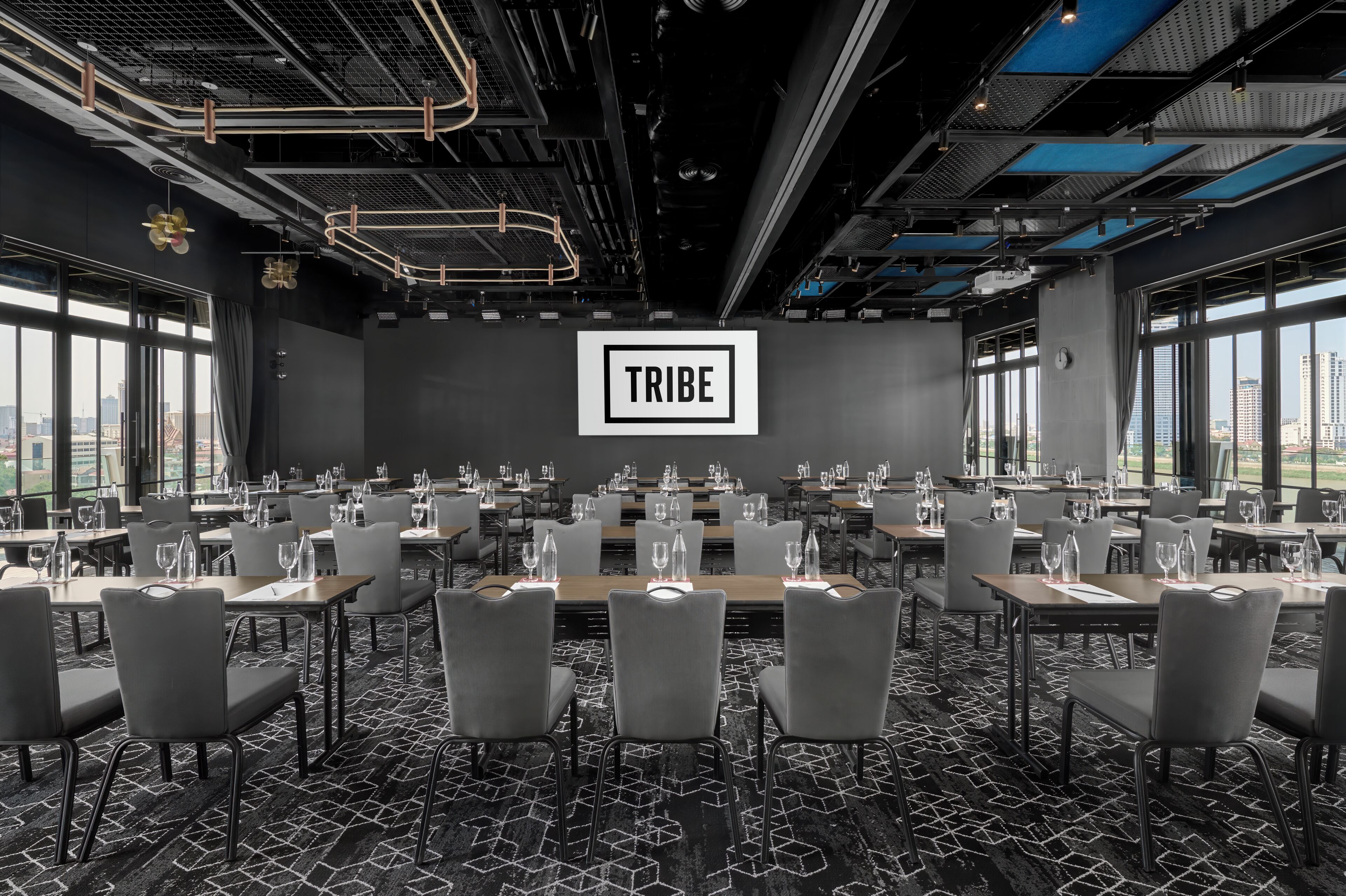 Una sala de conferencias con varias mesas y sillas, un cartel grande con la palabra "TRIBE" y ventanas amplias con vistas a la ciudad.