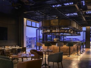 A modern bar area with a city view, bar stools, and tables with glasses. - TRIBE Phnom Penh Post Office Square (Phnom Penh)
