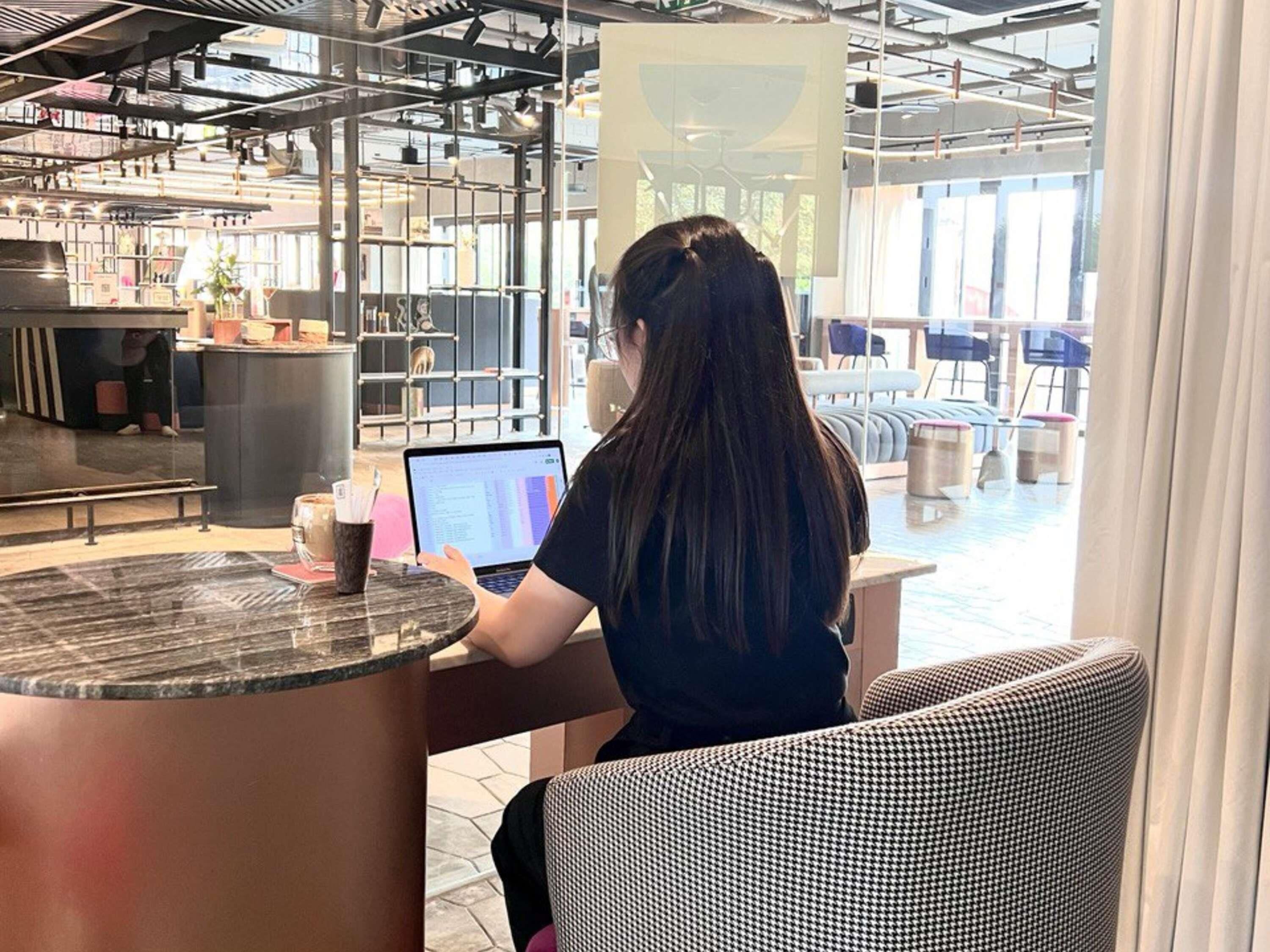 A person working on a laptop at a table with a view of a pool and outdoor seating.