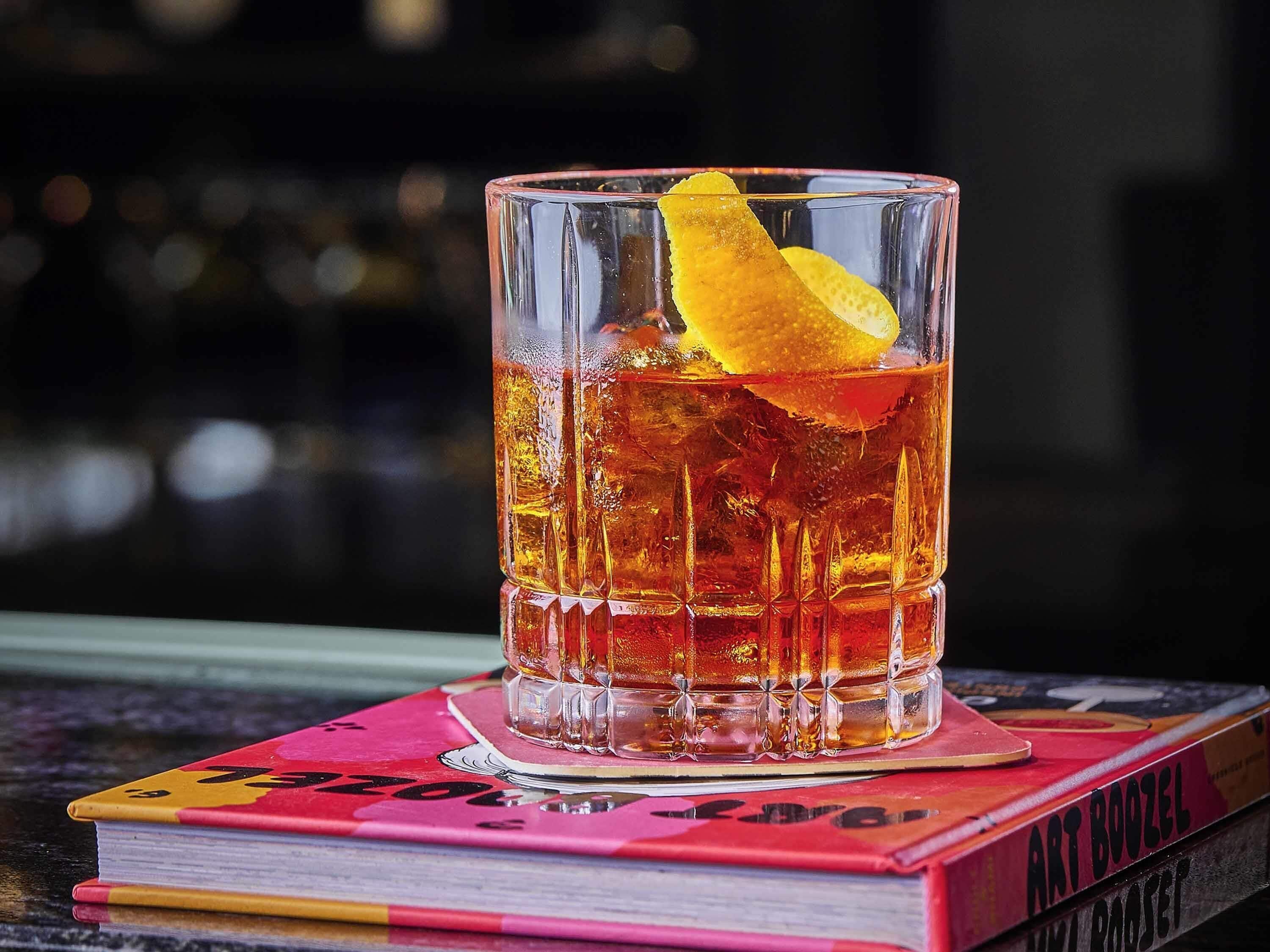 A glass of amber-colored beverage with a lemon slice garnish on a coaster, placed on a stack of books.