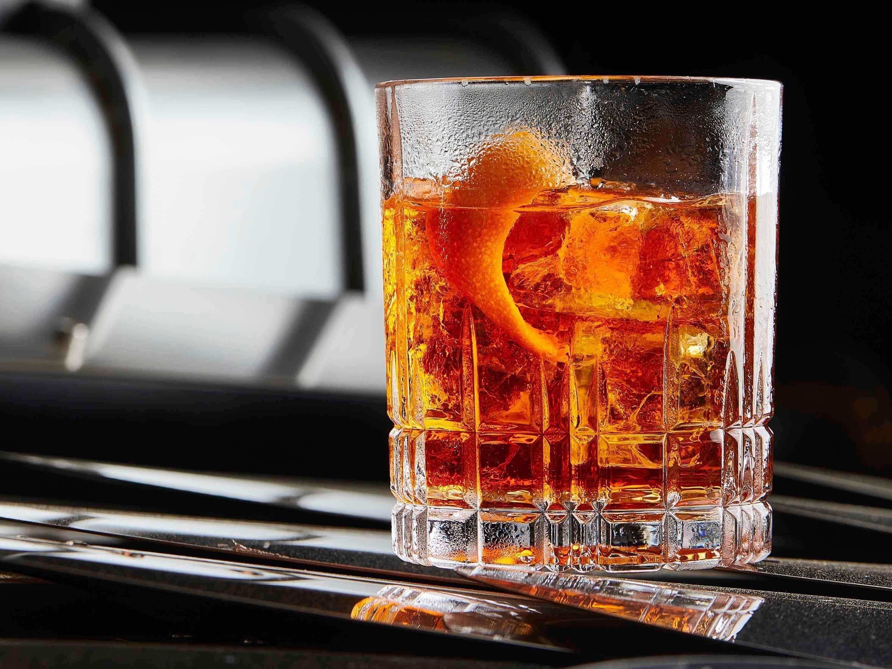 Un vaso transparente con un líquido ámbar oscuro, cubitos de hielo y un adorno cítrico sobre una superficie reflectante.