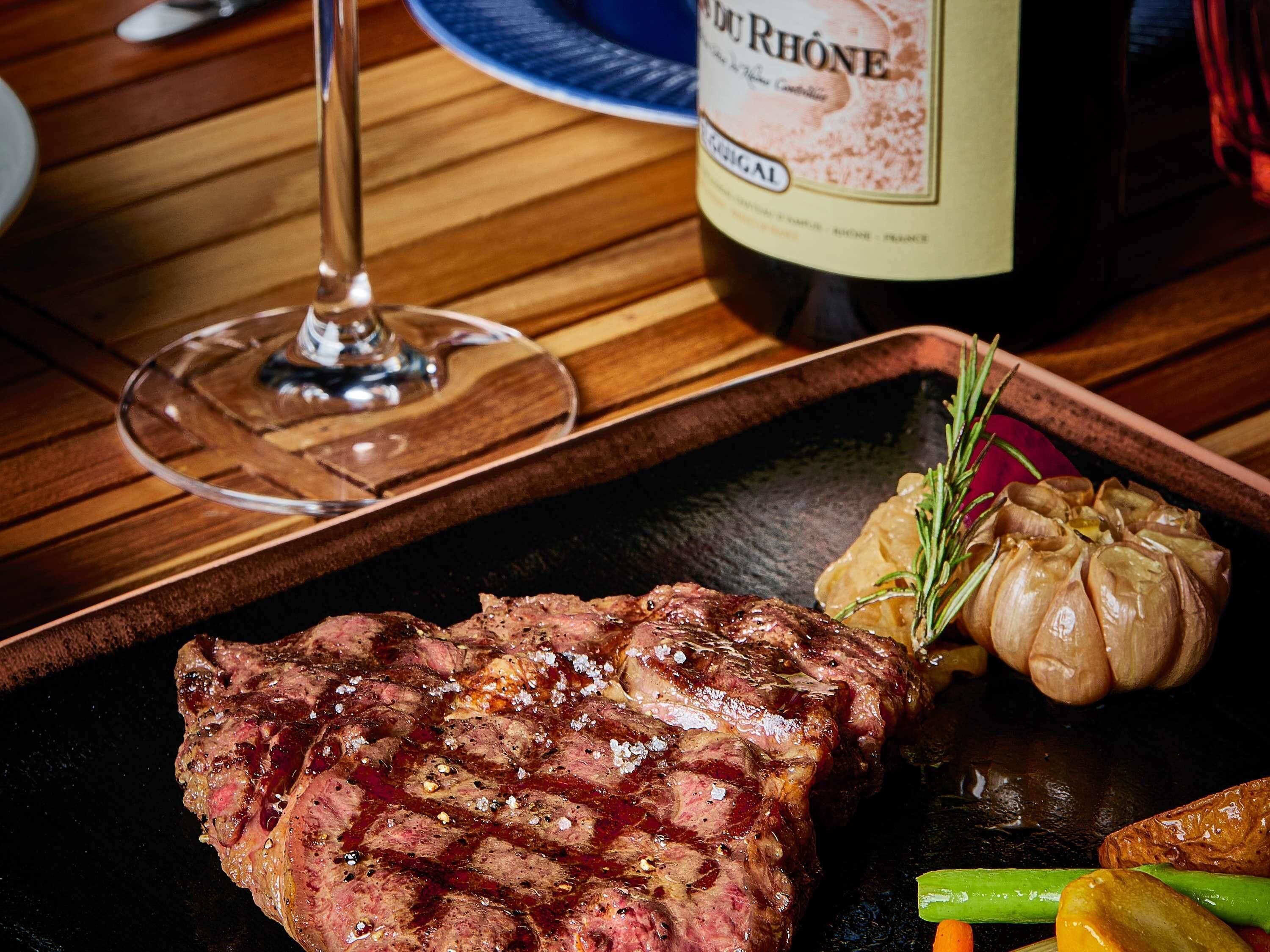 A steak, garlic, and vegetables on a plate with a bottle of wine in the background.