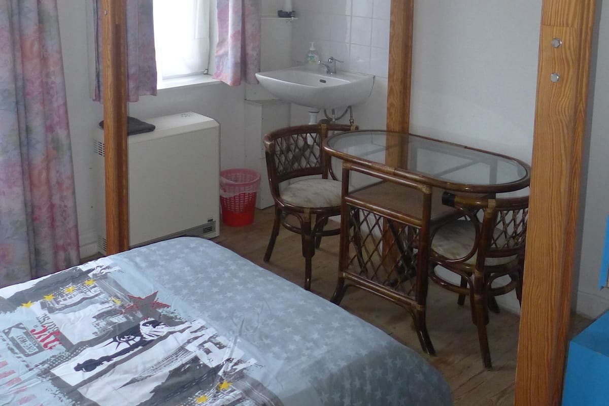 Un lit superposé avec un bureau et une chaise, un lit recouvert d’une couverture à motifs, une fenêtre avec des rideaux et un lavabo dans un coin.