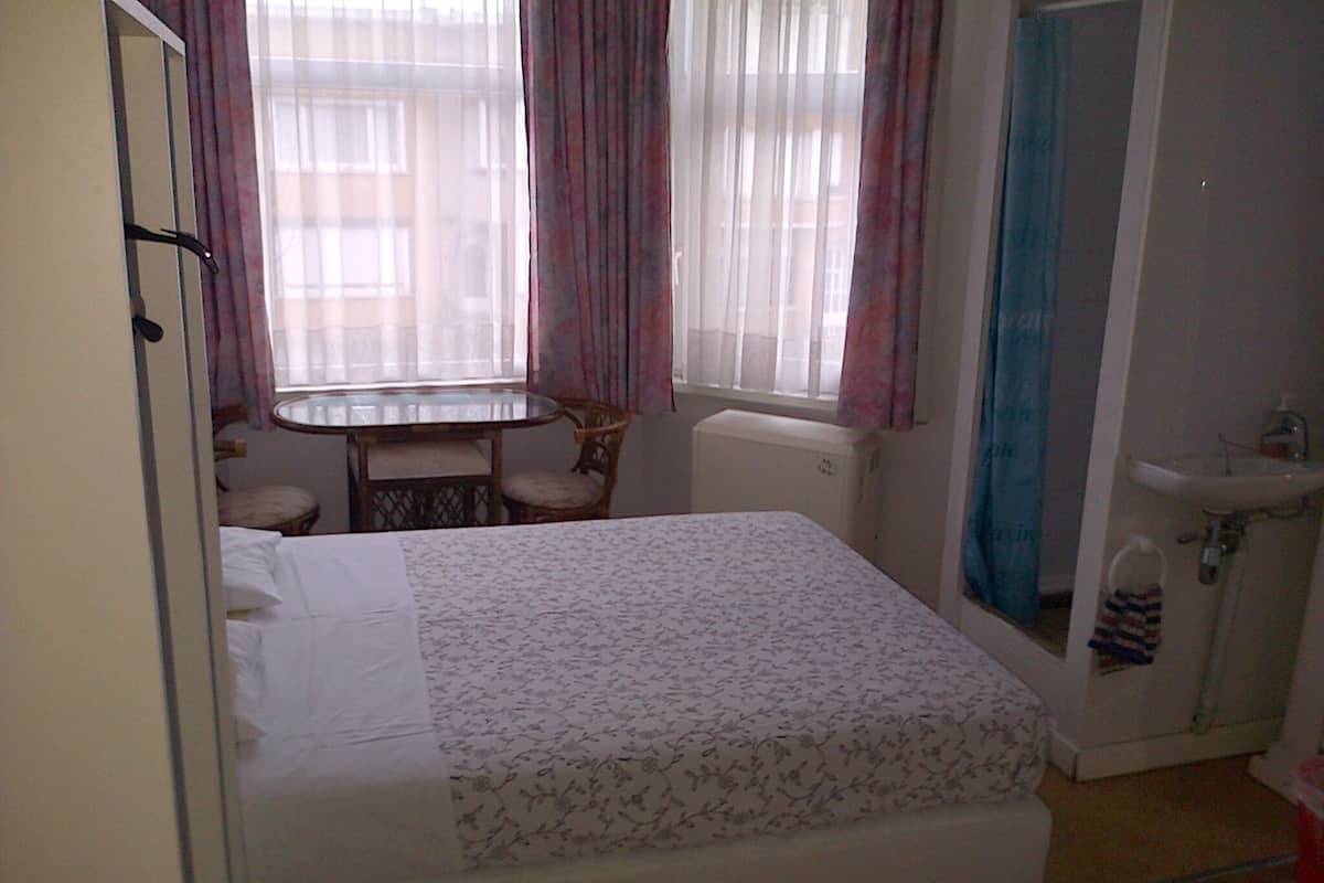 Une chambre à coucher comprenant un lit, une chaise, une petite table, une fenêtre avec des rideaux et une salle de bain avec un lavabo.