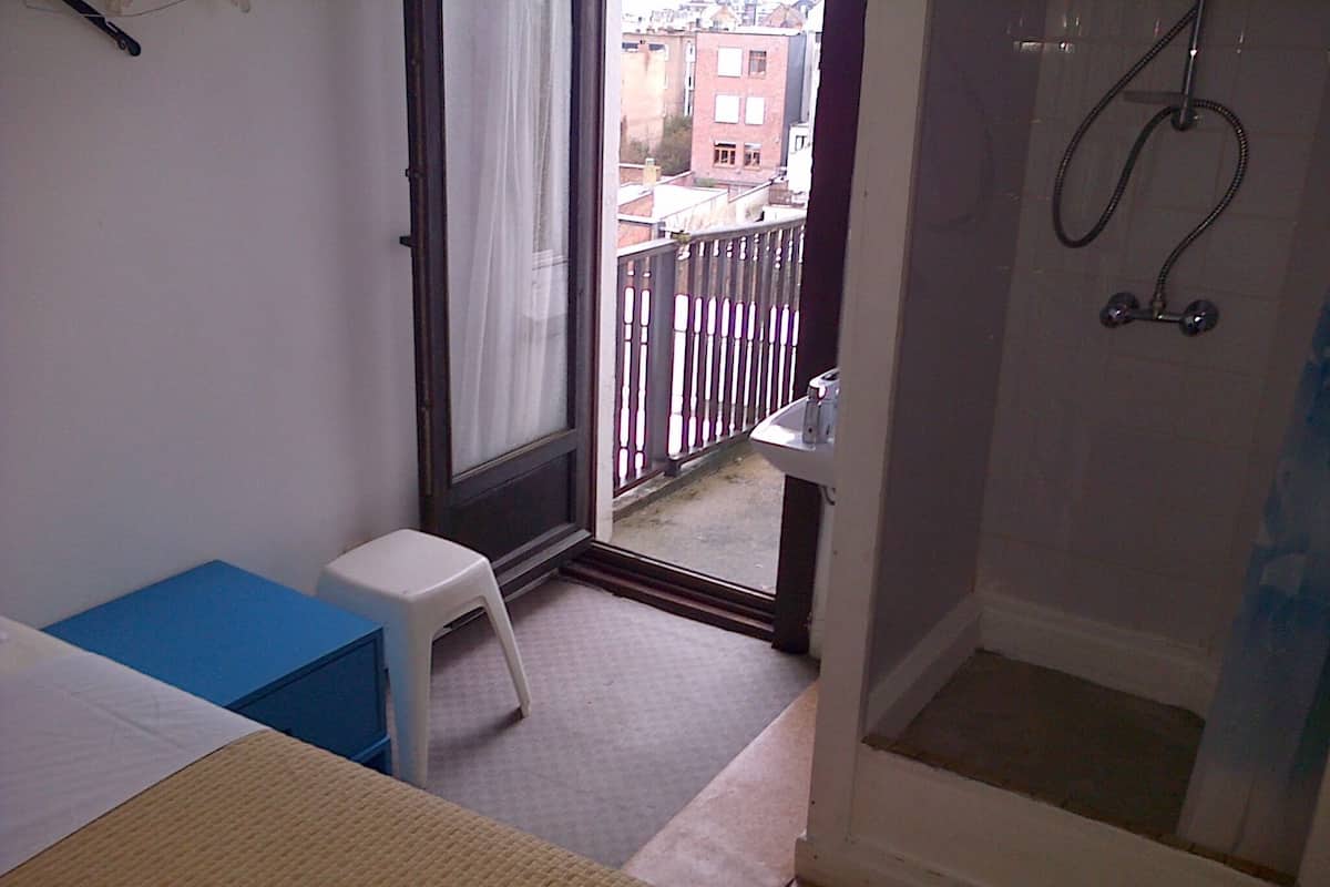 Une petite pièce, de taille modeste, comprenant un lit, un tabouret blanc, un espace douche et une fenêtre donnant sur la ville.
