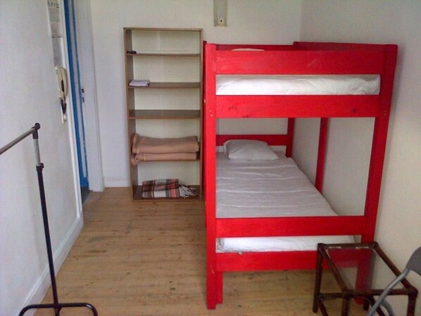 A bunk bed with a red frame, a wooden floor, a metal chair, and a shelf unit with folded blankets.