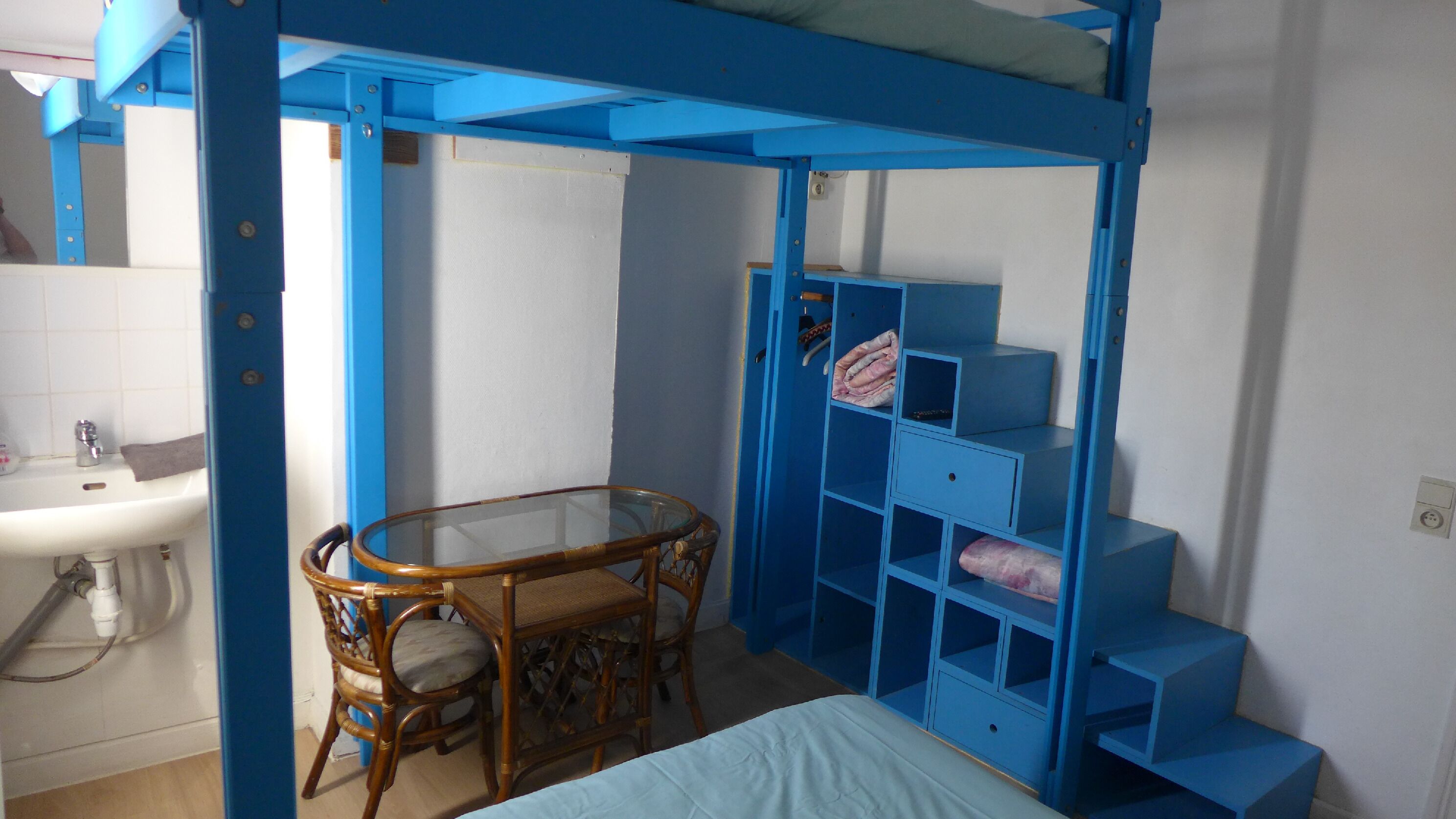 Quarto com uma cama de solteiro, uma mesinha com cadeiras, pia e uma estante azul.
