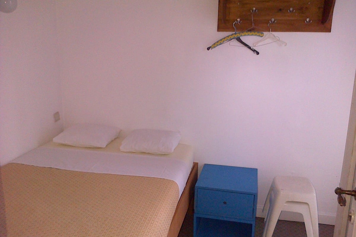 Une chambre à coucher comprenant un lit, une table de chevet, un petit tabouret et une étagère en bois avec des cintres.