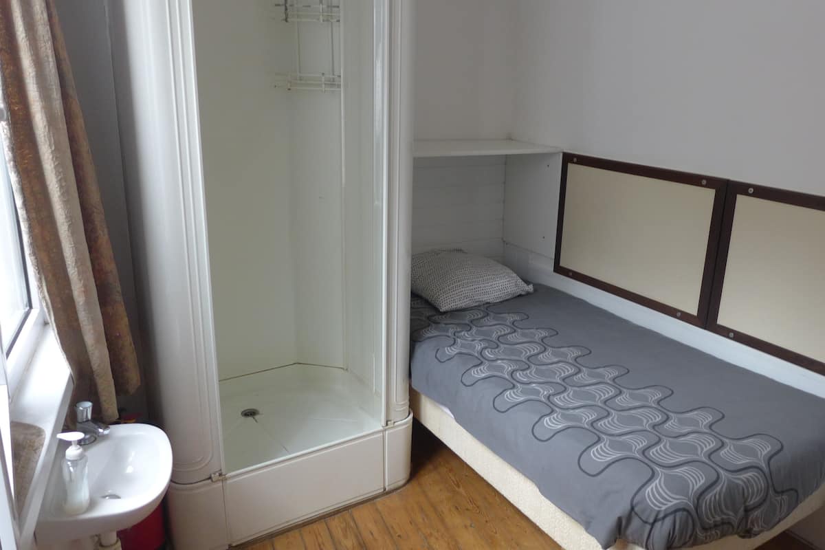 Une petite pièce, de taille modeste, comprenant une douche, un lit simple avec une couverture à motifs, une table de chevet et un lavabo.