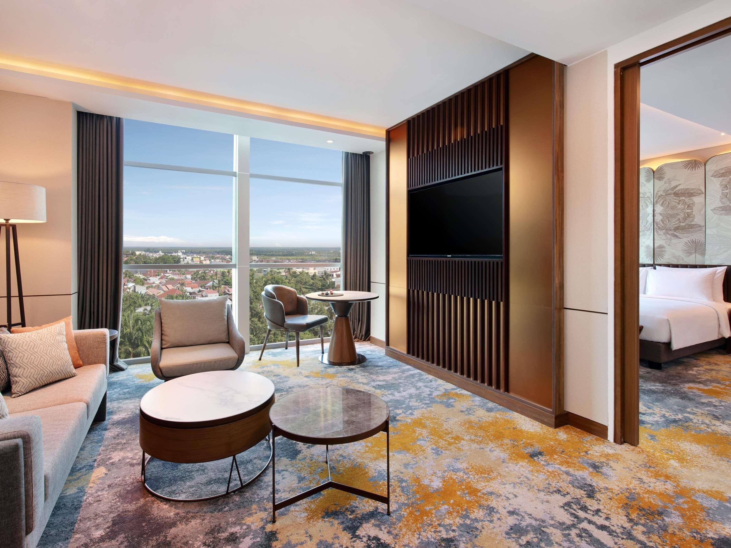 Quarto de hotel com sofá, poltrona, mesas redondas, televisão e uma grande janela com vista para a cidade.