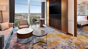 Une chambre d’hôtel équipée d’un canapé, d’un fauteuil, de petites tables rondes, d’une télévision et d’une grande fenêtre donnant sur la ville.