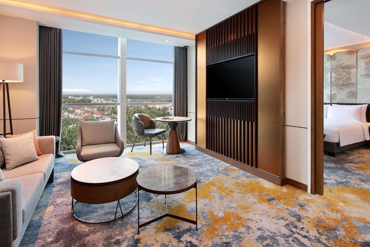 Une chambre d’hôtel équipée d’un canapé, d’un fauteuil, de petites tables rondes, d’une télévision et d  ’une grande fenêtre donnant sur la ville.