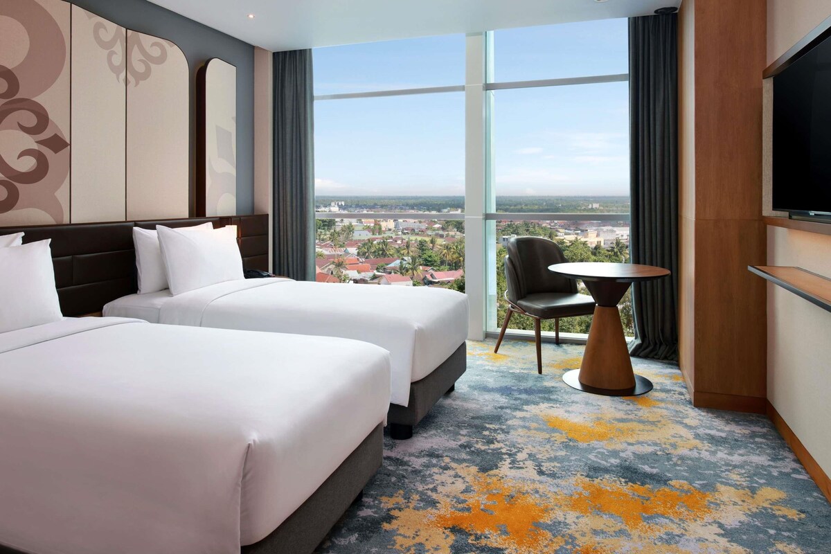 A hotel room with two beds, a TV, a small table, and a view of a cityscape.