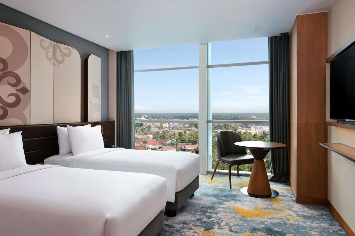 A hotel room with two beds, a TV, a small table, and a view of a cityscape.
