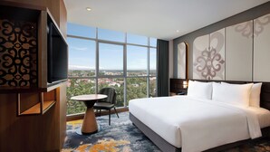 A hotel room with a large bed, a TV mounted on the wall, a small round table, a chair, and a view of the city through large windows. - Mercure Pangkalan BUN (Pangkalan Bun)