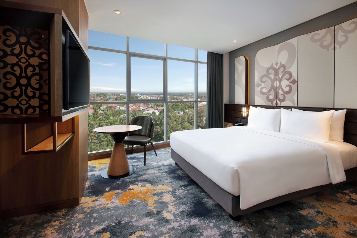A hotel room with a large bed, a TV mounted on the wall, a small round table, a chair, and a view of the city through large windows.