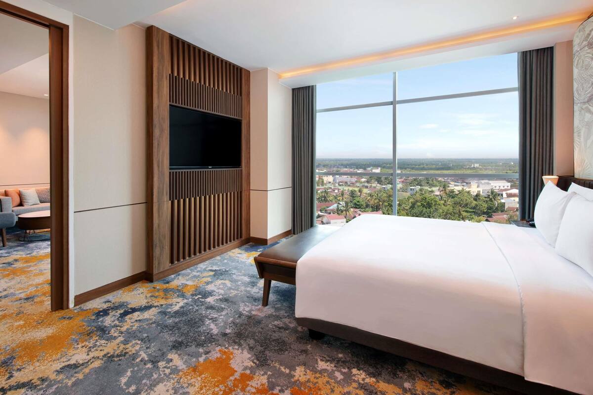 A hotel room with a large bed, a TV mounted on a wooden panel, a view of a cityscape, and a colorful carpet.