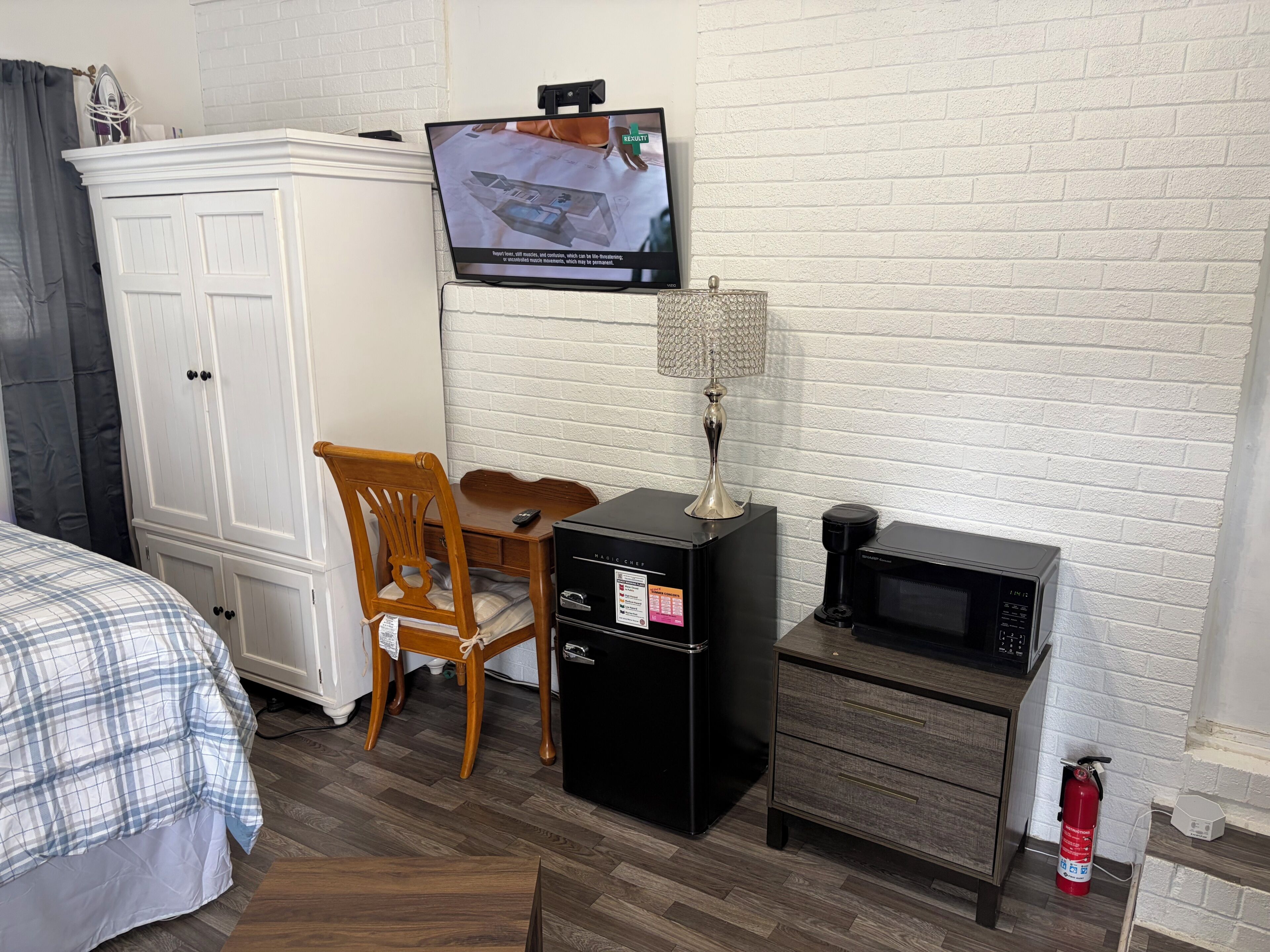 Quarto de hotel com uma cama, guarda-roupa, televisão, escrivaninha com cadeira, máquina de café, micro-ondas e extintor de incêndio.