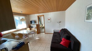 Ein Wohnzimmer mit schwarzem Sofa, einem Esstisch mit Stühlen, einem Fernseher und einem Fenster mit Vorhängen.