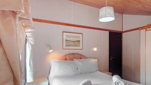Un lit bien fait, avec du linge de lit blanc et deux serviettes enroulées, une table de chevet avec une lampe et un tableau encadré au mur.