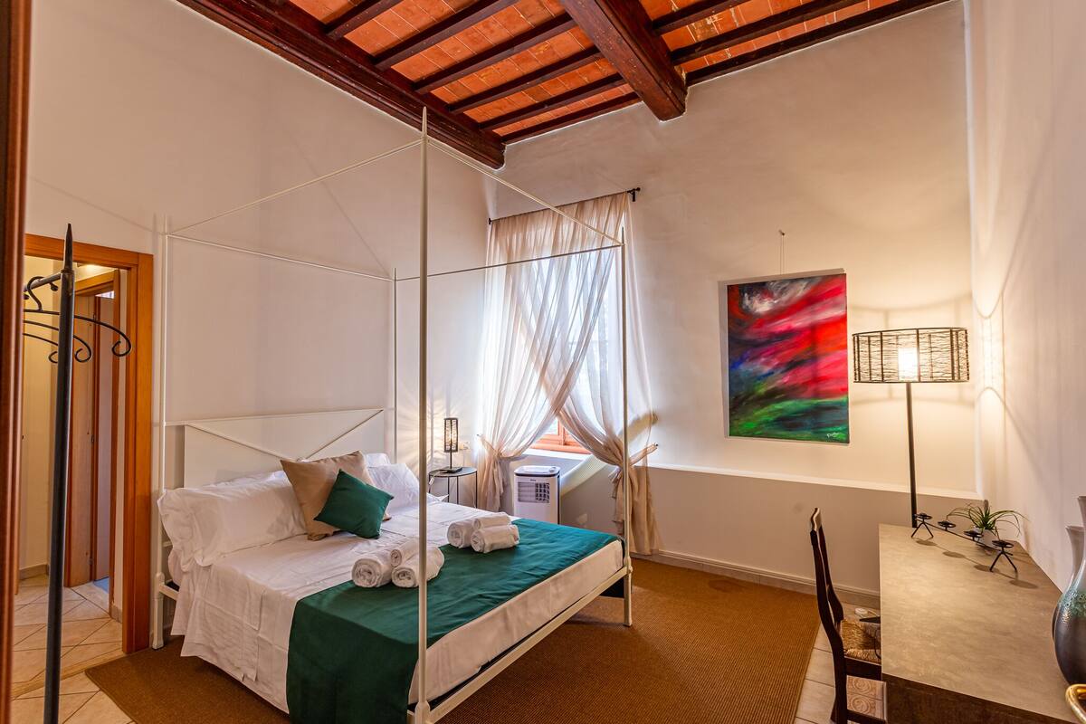 Una cama con dosel con sábanas blancas, una colcha verde y una ventana con cortinas traslúcidas.