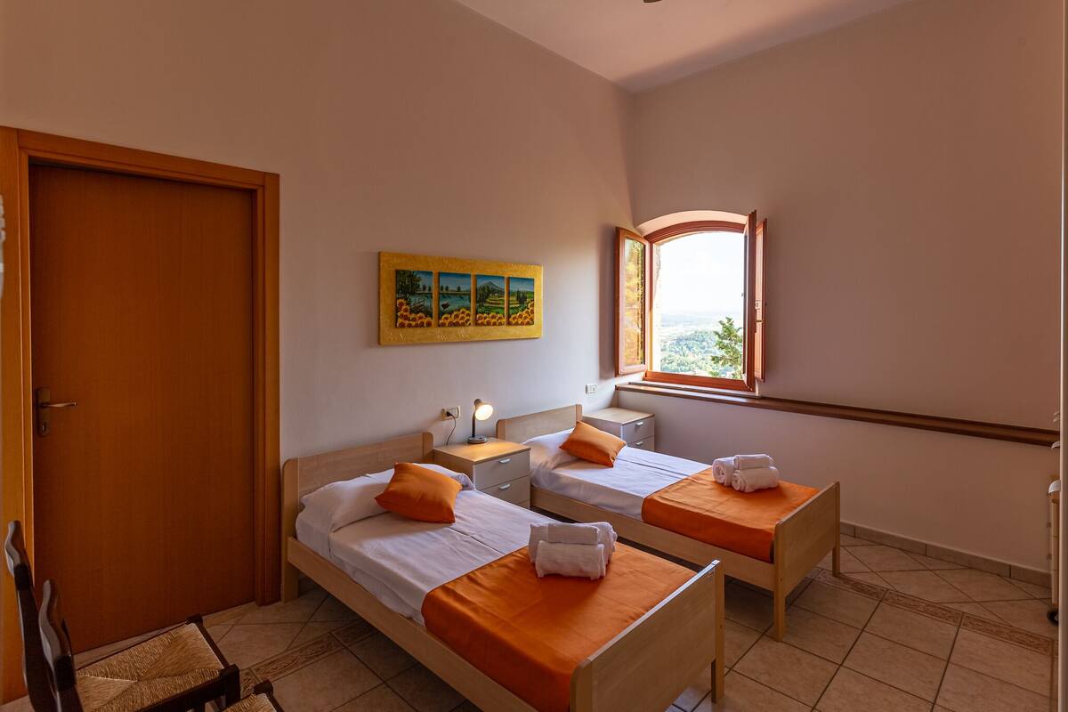 Habitación de hotel con dos camas individuales, una puerta de madera, una mesita con una lámpara, una ventana con vistas y un cuadro en la pared.